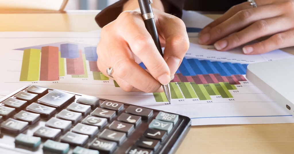 A close-up of a person's hand using a pen to analyze colorful data bar charts on paper next to a calculator.