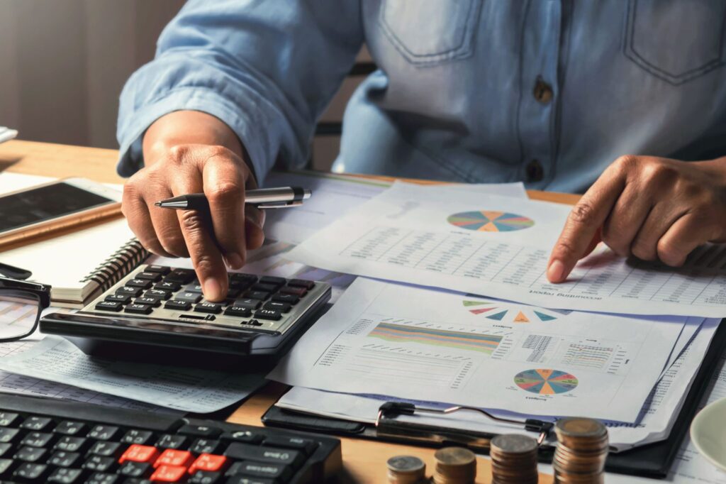 Hands using a calculator and reviewing financial reports with charts, coins, glasses, and a smartphone on a desk.