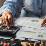 Hands using a calculator and reviewing financial reports with charts, coins, glasses, and a smartphone on a desk.