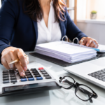 Bookkeeper using a calculator at a desk with files and a laptop.