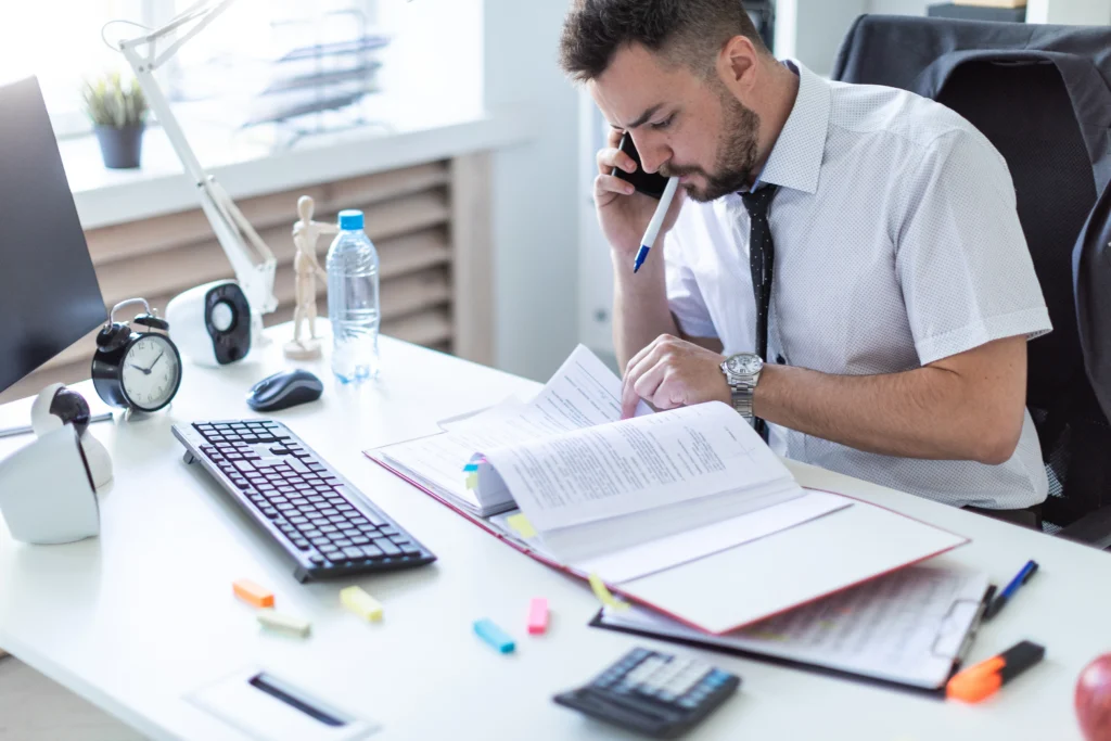 Stressed businessman talking on the phone while reviewing paperwork at his desk.