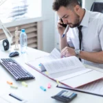 Stressed businessman talking on the phone while reviewing paperwork at his desk.