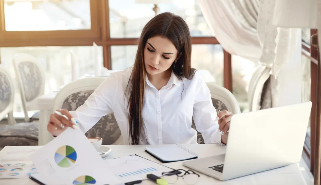 Businesswoman reviewing printed chart documents beside open laptop at desk.