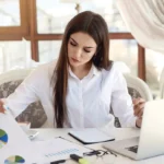 Businesswoman reviewing printed chart documents beside open laptop at desk.
