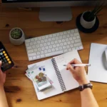 Top-down view of someone writing budget notes next to Euro cash and a phone calculator.